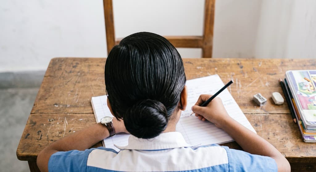Student writing in a notebook at a wooden desk